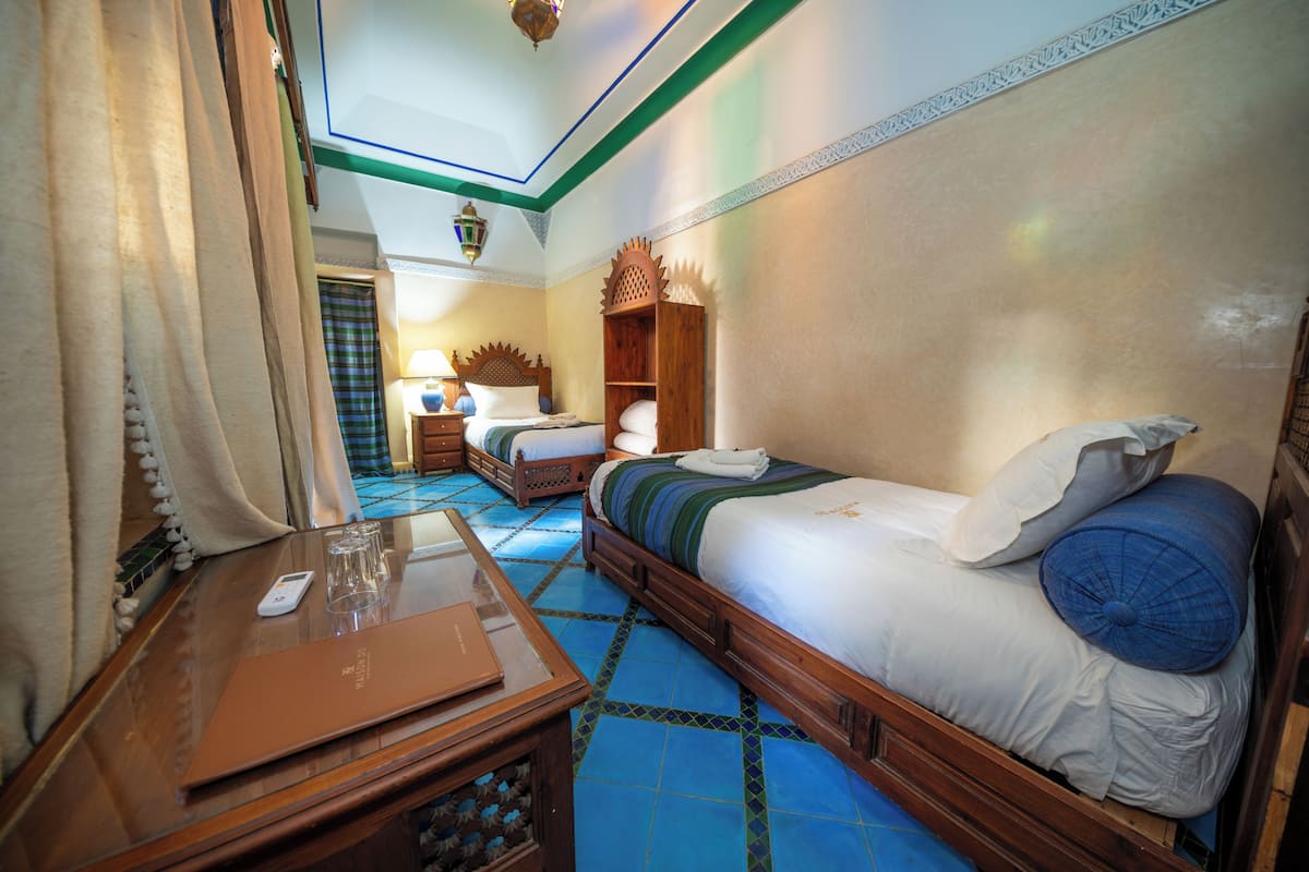 Une chambre d’hôtel avec un grand lit, une table de chevet en bois, un bureau avec un ordinateur portable et des lunettes, et un sol carrelé bleu.