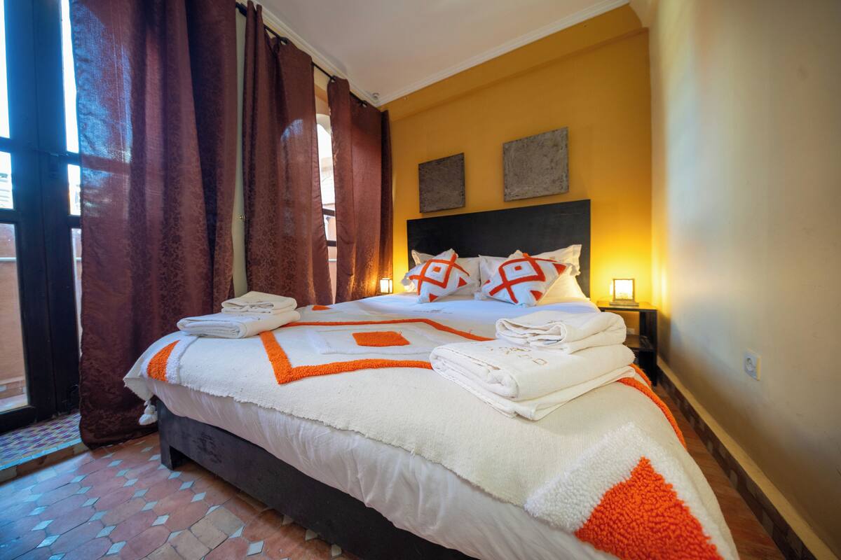 Un lit bien fait, avec une literie blanche et orange, une tête de lit et deux lampes de chevet.