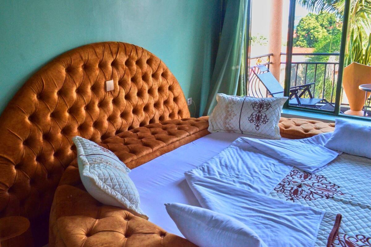 Un dormitorio con cabecera capitonada, una cama grande con ropa de cama blanca y vistas a un balcón con mobiliario exterior.