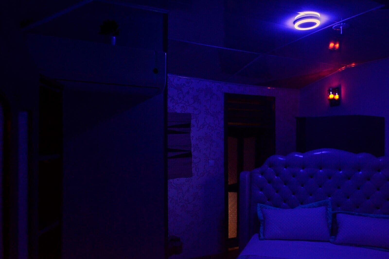 A dimly lit room with a tufted headboard bed, a visible door, and a patterned wall.