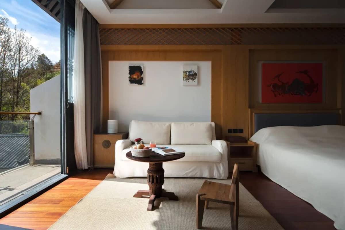 Ein modernes Hotelzimmer mit einem großen Bett, einem Sofa, einem kleinen runden Tisch mit einem Buch und einer Obstschale sowie Blick ins Freie.