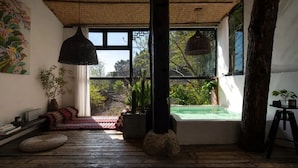 A room with a wooden floor, a large window with a view of greenery, a black hanging light, and a small table with a plant. - GRAND JOLI (Lijiang)