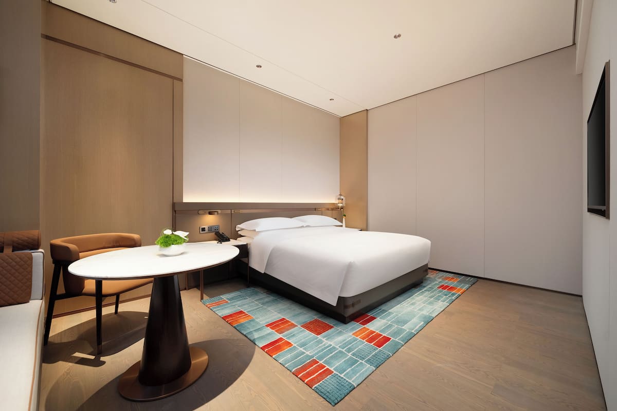Quarto de hotel moderno com uma cama bem arrumada, uma pequena mesa redonda com uma planta e uma cadeira.