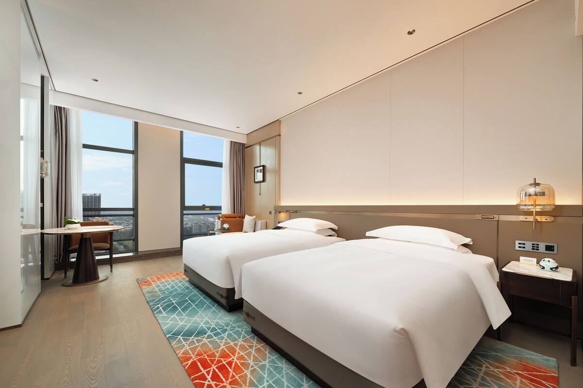 Quarto de hotel moderno com duas camas, uma escrivaninha, uma cadeira, mesa de cabeceira e uma janela ampla com vista para a cidade.