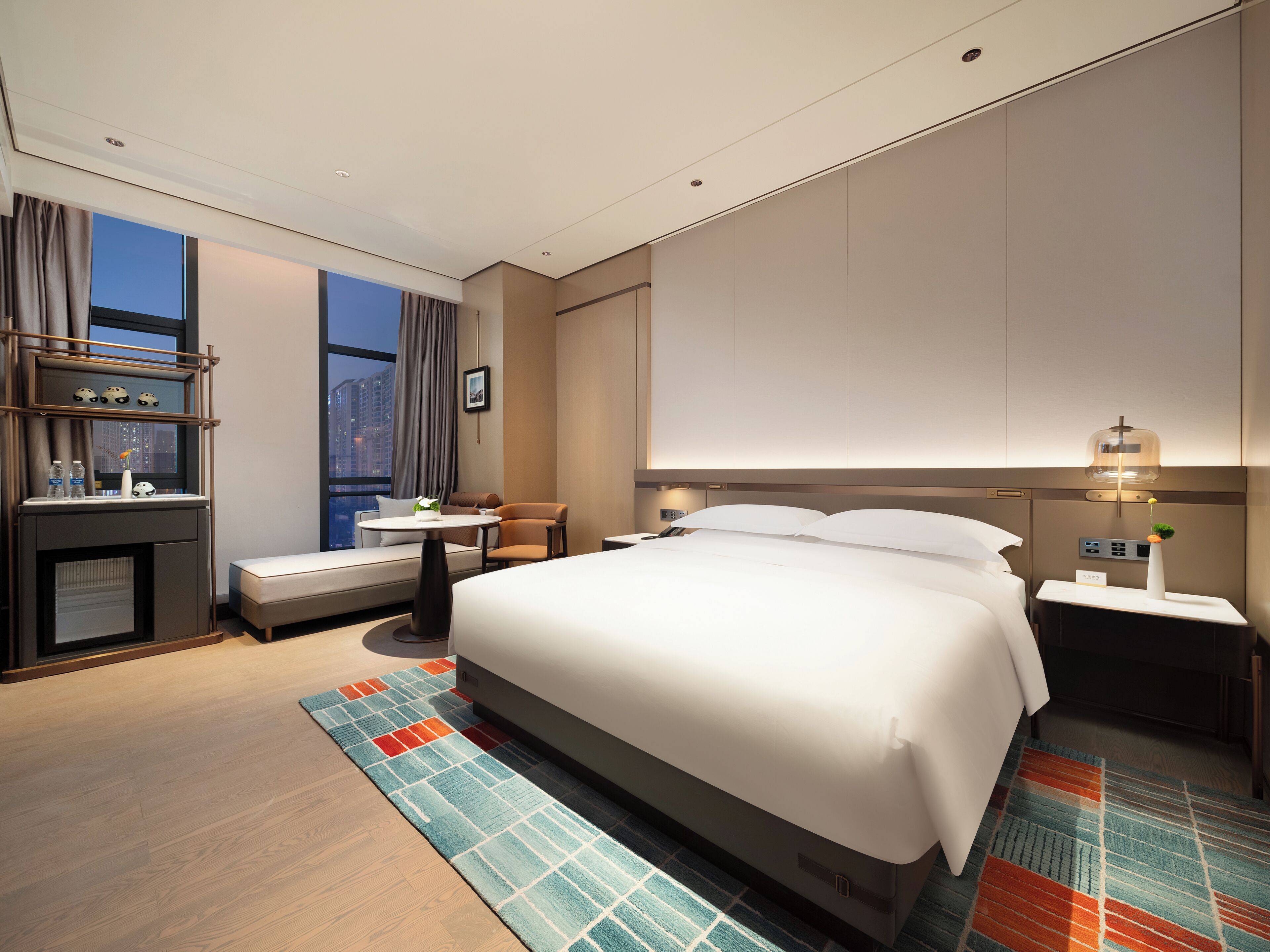 Ein modernes Hotelzimmer mit einem großen Bett, einem Schreibtisch mit Stuhl, einer kleinen Sitzecke und Blick auf die Stadtlandschaft.