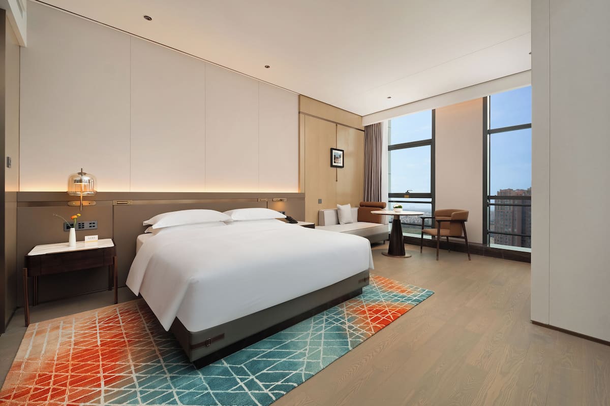 Quarto de hotel moderno com uma cama grande, uma mesinha, um sofá e uma varanda com vista para a cidade.