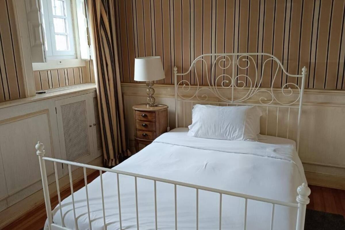Una cama bien tendida con estructura metálica blanca, una mesita de noche con lámpara y una ventana con cortinas.