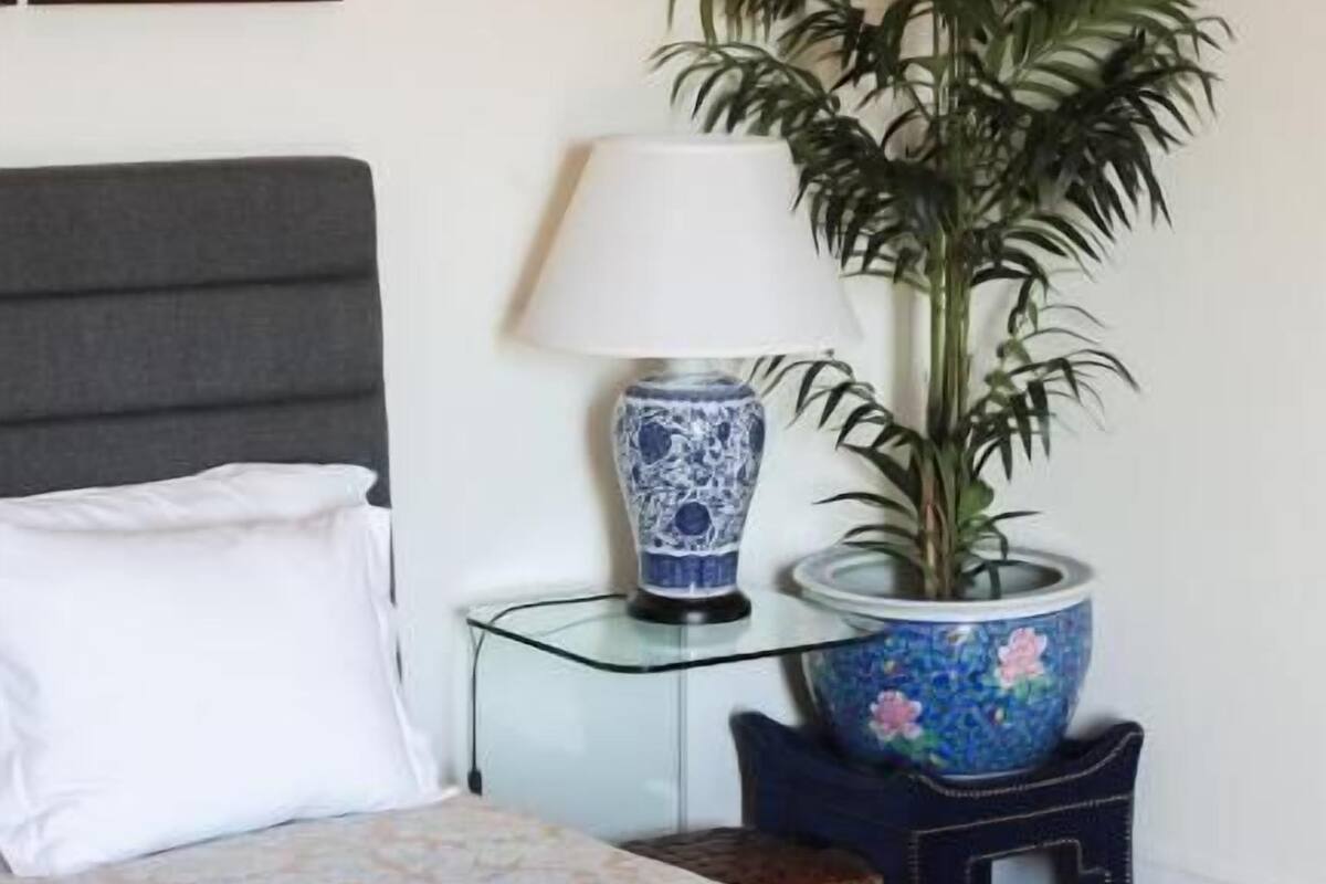 Una cama bien hecha con una colcha estampada, un cabecero gris, un cuadro enmarcado en la pared, un jarrón azul y blanco sobre una mesa de vidrio y una planta en maceta.