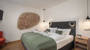 A modern bedroom with a brick wall feature, a neatly made bed with white linens, a green throw blanket, and a wooden nightstand with a lamp. - dreamcation Altstadthotel Regensburg (Regensburg)