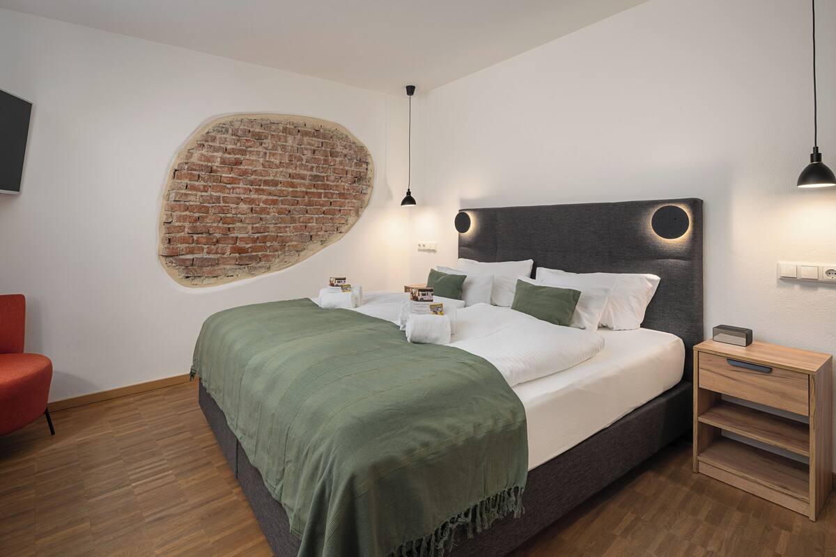 Une chambre moderne avec un mur en briques apparentes, un lit bien fait avec du linge de lit blanc, un plaid vert et une table de chevet en bois avec une lampe.