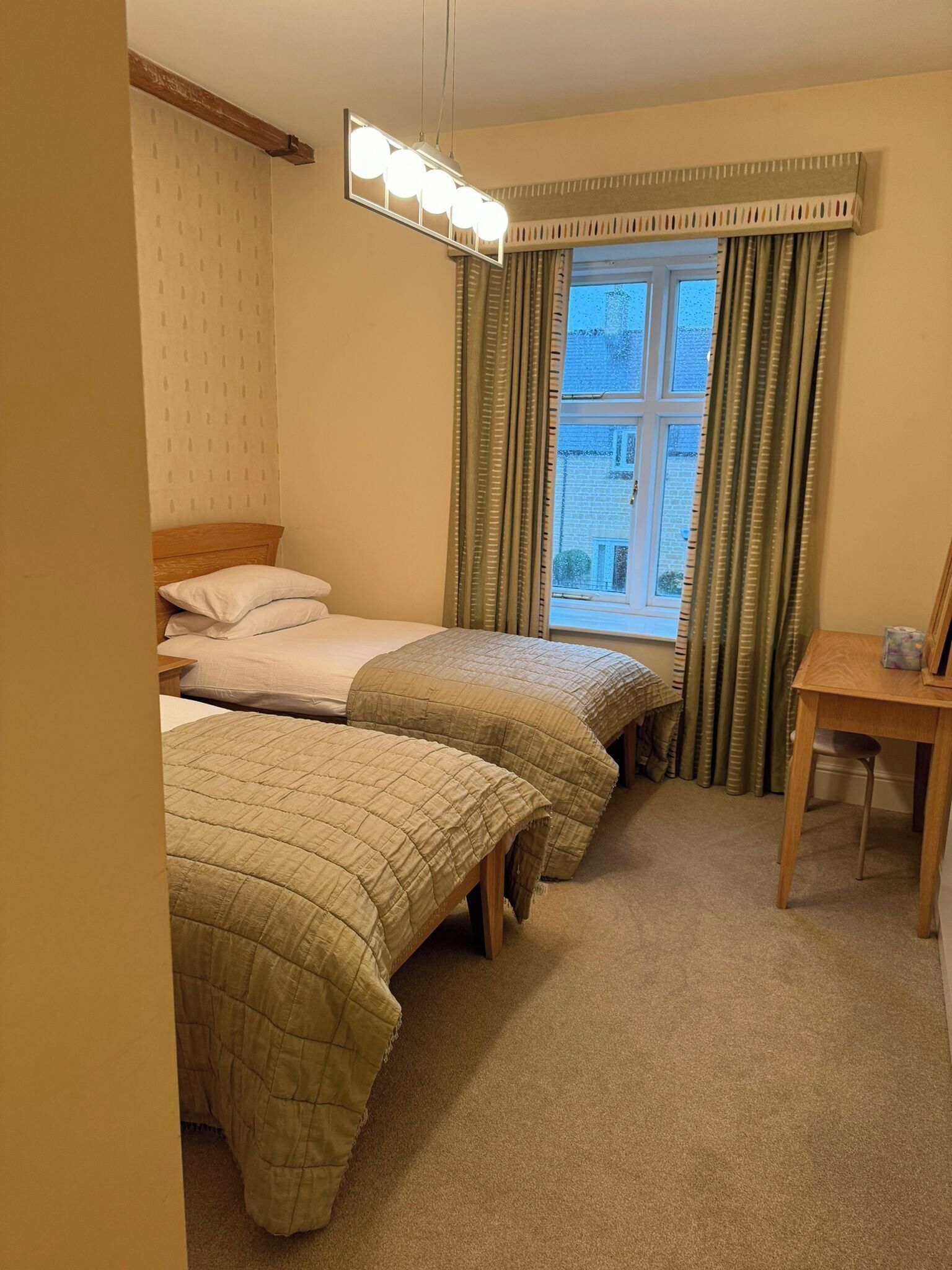 A hotel room with two beds, a small table, a chair, a window with curtains, and a ceiling light fixture.