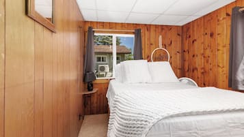 Ein Schlafzimmer mit Holzwänden, einem Bett mit weißer Bettwäsche, einem Fenster mit Vorhängen und einem kleinen Nachttisch mit einer Lampe.