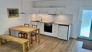 Una cucina compatta con mobili bianchi, forno integrato e lavastoviglie. C'è un tavolo da pranzo in legno con sedie e una panca.