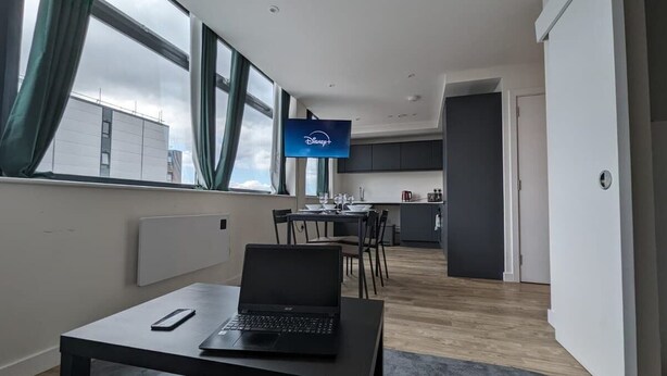 A modern room with a laptop on a table, a flat-screen TV mounted on the wall, a dining area with chairs, and a kitchen in the background.