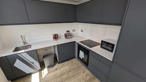 A compact kitchen with grey cabinets, a white countertop, a built-in microwave, and a kettle.