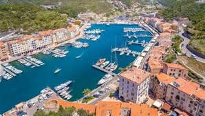 An aerial view of a coastal town with a marina, buildings with red-tiled roofs, and a hilly landscape in the background.