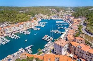 Vista aérea de un pueblo costero con un puerto deportivo, edificios con tejados rojos y un paisaje montañoso al fondo.
