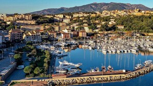 A marina with boats docked, a rocky breakwater, and a town with buildings and mountains in the background.
