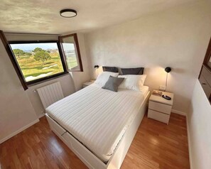 A bedroom with a bed, a nightstand, a lamp, a window with a view of a green landscape, and a radiator. - Par71 - Marco Simone golf club Roma (Marco Simone)