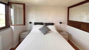 Ein Einzelbett mit weißer Matratze und grauem Kopfkissen, an beiden Seiten jeweils ein Nachttisch mit einer Lampe, in einem Zimmer mit Fenster und schlichter Wand.