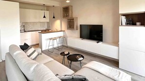 A modern living space with a sofa, a coffee table, a TV, and a kitchen area. - Par71 - Marco Simone golf club Roma (Marco Simone)