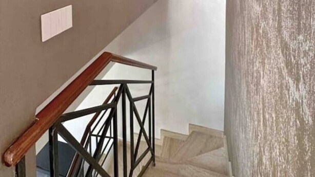 Eine Treppe mit Holzbalkon und Metallspindeln, die zu einer Zwischenebene mit einer weißen Wand und einer strukturierten Wand rechts führt.