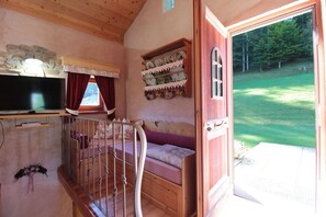 A cozy room with a television, a stone wall, a window with curtains, a shelf with dishes, and a door leading outside.