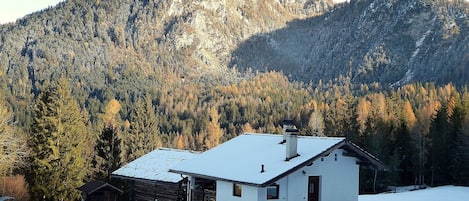 A white house with a snow-covered roof, situated in a snowy landscape with a forested mountain in the background.