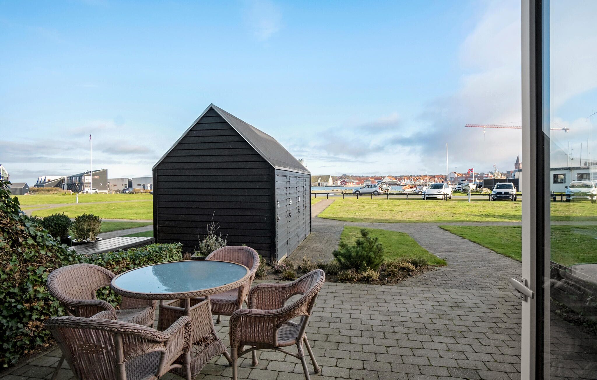 A patio area with wicker furniture and a glass table, a black shed, and a view of a marina with parked cars and boats.