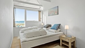 Une chambre à coucher avec un lit, une table de chevet, une lampe, une fenêtre donnant sur la mer et un tableau accroché au mur.