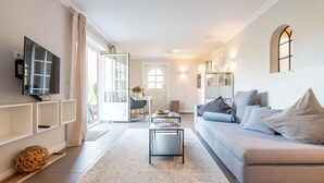 A modern living room with a sofa, coffee table, and TV. There is a door leading to a balcony, and a small table with a vase of flowers.