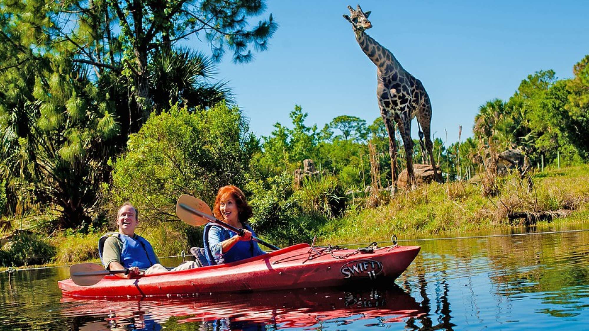 Zwei Personen paddeln in einem ruhigen Fluss, umgeben von üppigem Grün, mit einer Giraffe im Hintergrund.