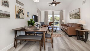 Una sala de estar moderna con comedor, que presenta un sofá de cuero marrón, una mesa redonda de madera y una mesa de comedor con platos, vasos y un jarrón de flores.