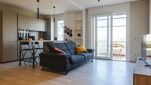 A modern living room with a sofa, a coffee table, and a TV stand. There is a kitchen area with a dining table and chairs. The room has large windows and a balcony with outdoor furniture. - Cinecitt Dreams in Rome (Rome)