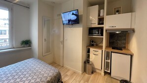Una habitación compacta con cama, televisor montado en la pared, una kitchenette pequeña con microondas y un armario.