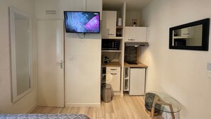 Una habitación compacta con cama, televisión montada en un mueble, kitchenette pequeña y un espejo en la pared.