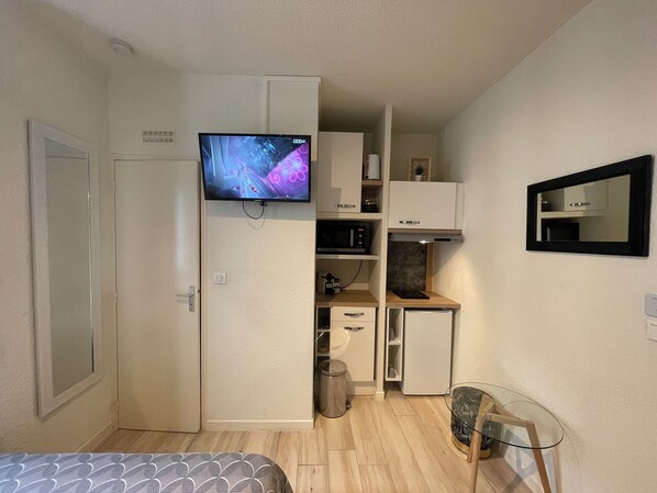 A compact room with a bed, a TV mounted on a cabinet, a small kitchenette, and a mirror on the wall.