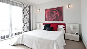 A modern hotel room with a large bed, a red rose artwork on the wall, and a nightstand with a lamp.