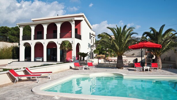 A pool area with a white building, red lounge chairs, and palm trees.