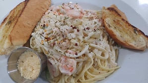 A plate of pasta with shrimp, garnished with herbs and red pepper flakes, accompanied by toasted bread slices and a small bowl of grated cheese. - Southern Corner Motel & Restaurant (Independence and Mango Creek)