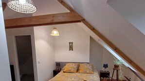 Een slaapkamer met een bed, houten balken, een hanglamp, een kleine tafel met een lamp en een potplant.