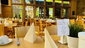 Ein Restaurant mit Holztischen und -stühlen, einem Menüständer und großen Fenstern mit Blick auf Gebäude und Grünflächen.