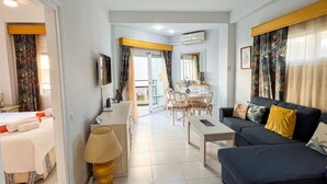 A hotel room with a bed, a sofa, a dining table with chairs, a TV, and a balcony with curtains.