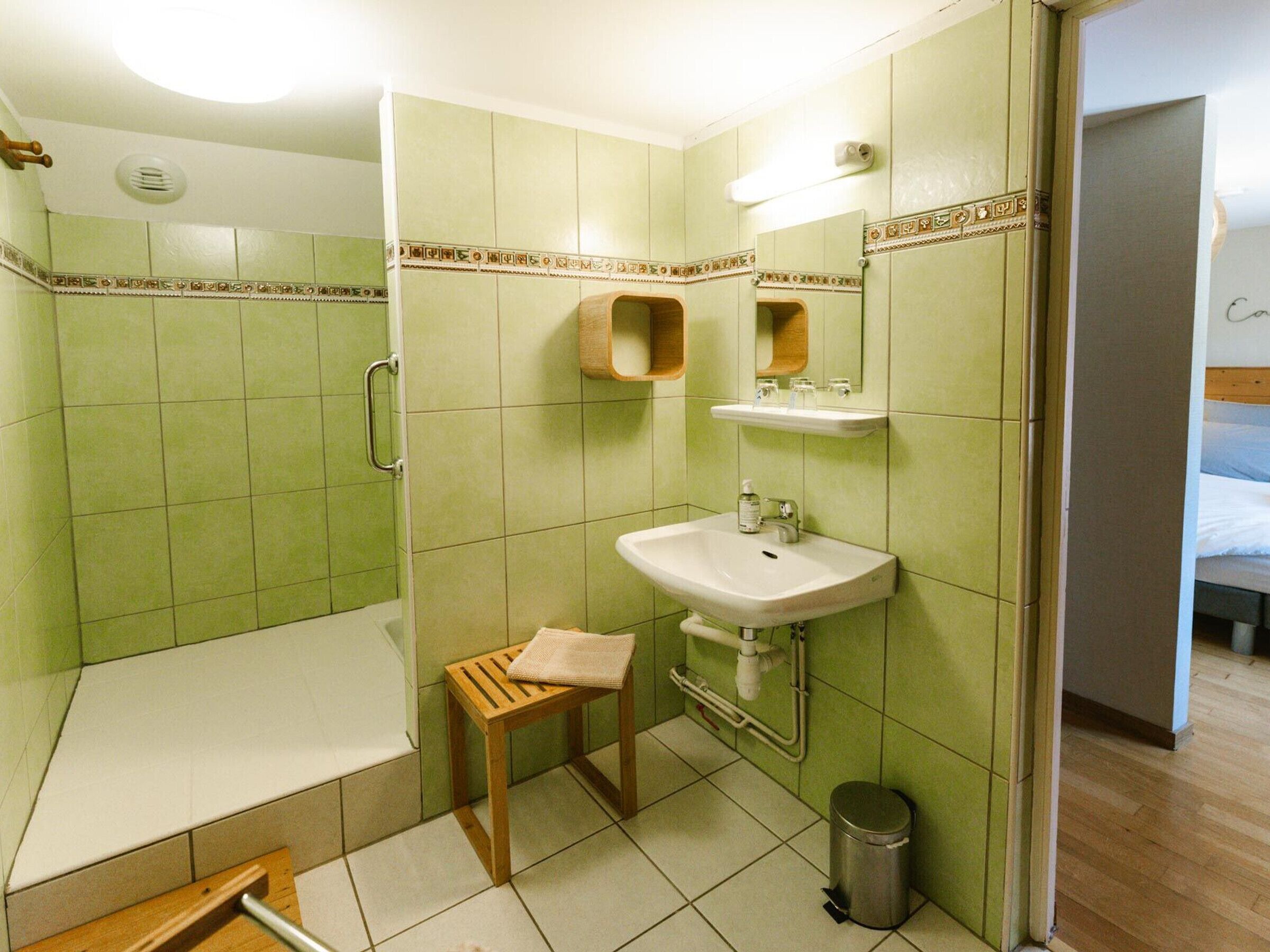 Un baño con baldosas verdes, un lavabo, un espejo, una ducha y un taburete de madera.