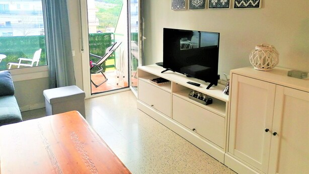 Un salon avec un téléviseur, un meuble blanc, une table en bois et un balcon avec une chaise.