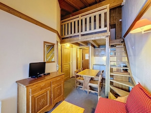 Ein kleines Zimmer mit einem Holztisch, einem Fernseher auf einer Holzkommode und einer Küchenzeile mit Mikrowelle und kleinem Kühlschrank.