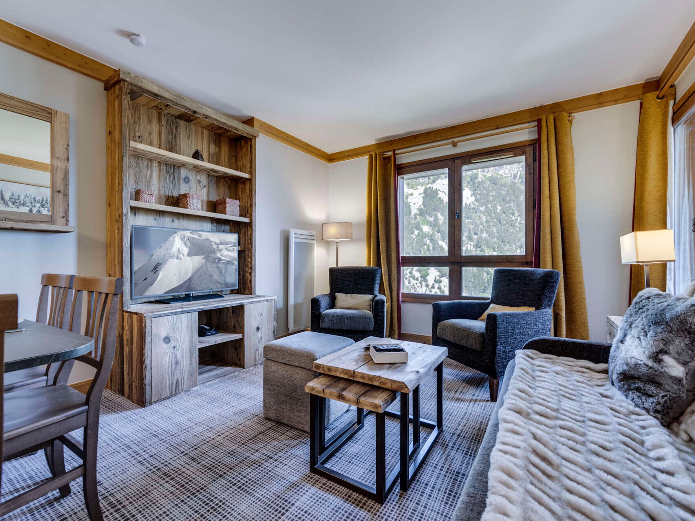 Ein gemütliches Zimmer mit einer Holztv-Konsole, einer Couch, zwei Sesseln, einem Esstisch mit Stühlen und einem großen Fenster mit Vorhängen.