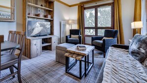 Une pièce chaleureuse avec un meuble TV en bois, un canapé, deux fauteuils, une table à manger avec des chaises et une grande fenêtre avec des rideaux.