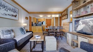 Un salon chaleureux avec un canapé, une table basse et un téléviseur fixé à un mur en bois. On y trouve également un coin cuisine avec des placards en bois et une table à manger avec des chaises.
