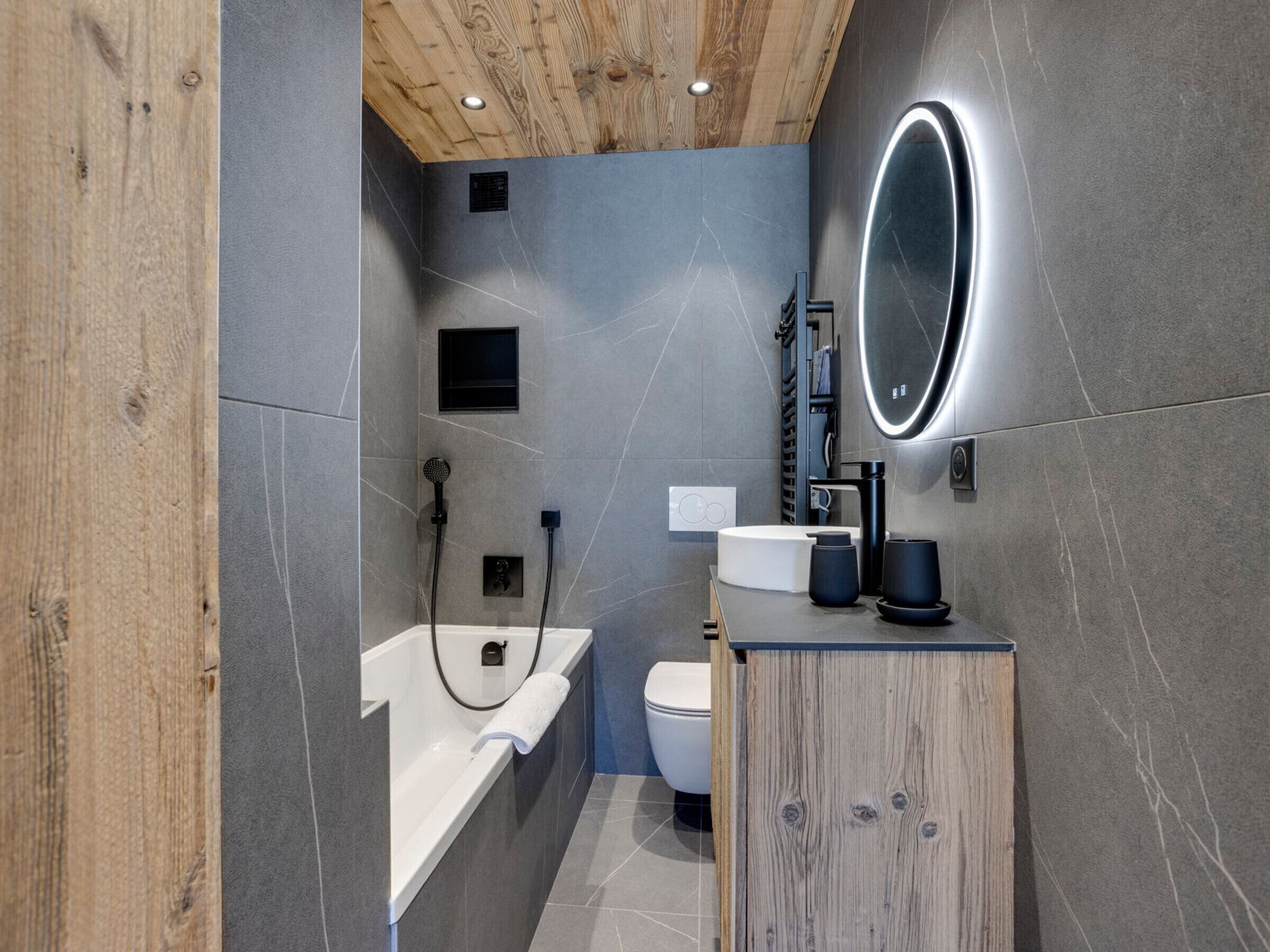 Et moderne badeværelse med træloft, grå flisemure, et hvidt badekar, et toilet og et rundt spejl med lys.